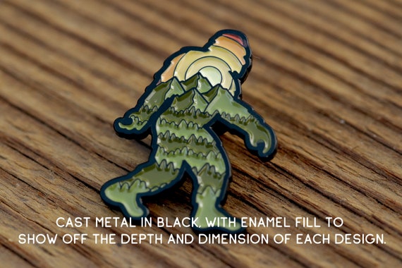 Angled on weathered wood, the Bigfoot Adventure Pin has text noting cast metal in black with enamel fill.