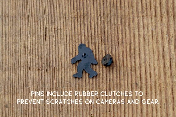 Back side of the Bigfoot Adventure Pin lies on wood with its rubber clutch and a note about scratches.