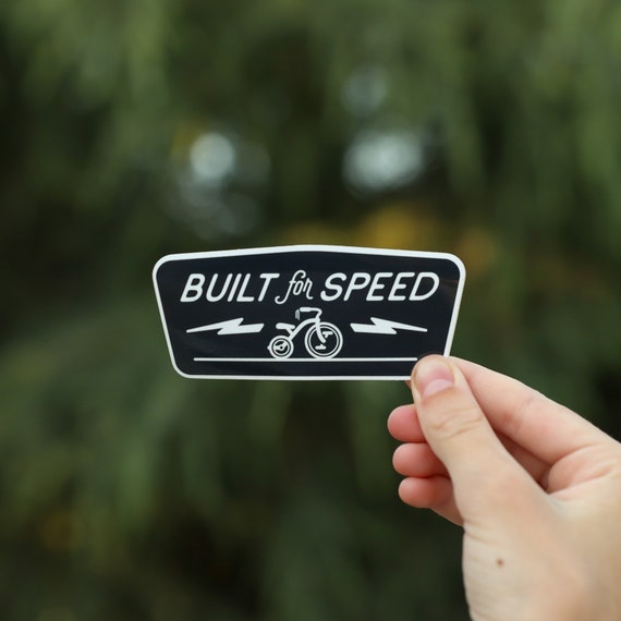 A hand holds the black Built for Speed sticker with its tricycle and lightning bolts against blurred green trees.