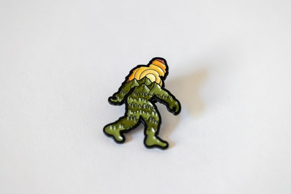 Plain white background lets the Bigfoot Adventure Pin show its sunset stripes and forested green legs.