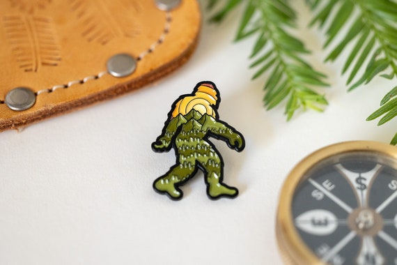 Beside a leather case and brass compass, the Bigfoot Adventure Pin rests on white with pine sprigs above.