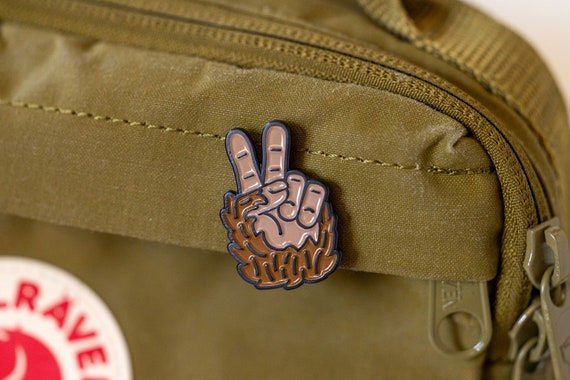 Clipped to the zippered pocket of an olive daypack, the Bigfoot Peace Pin sits above a round patch.