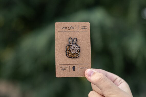 A hand holds up the kraft backing card in front of blurred green leaves, the Bigfoot Peace Pin centered.