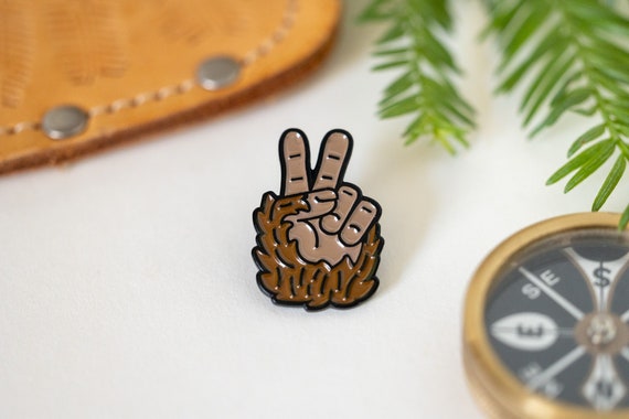 Beside a leather case and a brass compass, the Bigfoot Peace Pin rests on white with pine sprigs nearby.