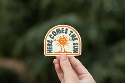 Fingers hold the arched Here Comes The Sun sticker with its smiling orange sun against blurred green trees.