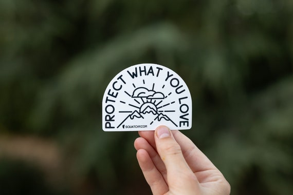 A hand holds the arched Protect What You Love sticker with its line drawn mountains against blurred green trees.