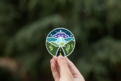 A hand holds up the round Roswell Conspiracy sticker, its UFO beaming over a desert road, against blurred green pines.