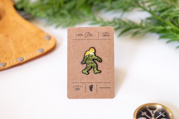 The Bigfoot Adventure Pin stays on its kraft lapel pin card between leather, greenery and a compass edge.