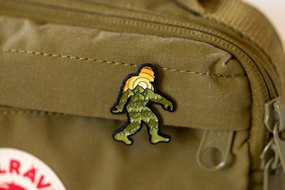 Fastened to an olive canvas pack near the zipper, the Bigfoot Adventure Pin sits above a red logo patch.