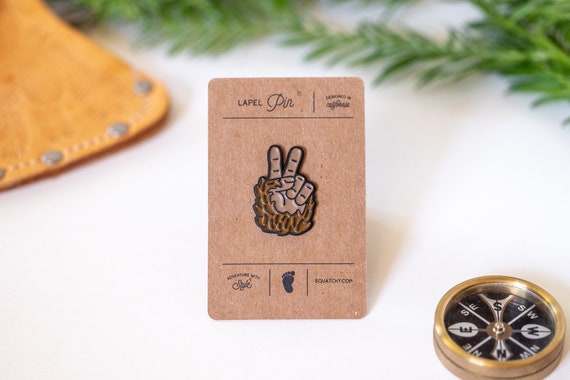 Mounted on its kraft lapel card, the Bigfoot Peace Pin lies flat between a leather flap and a compass.