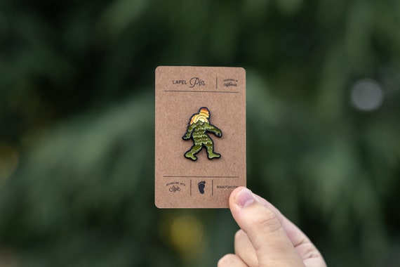 A hand holds the carded Bigfoot Adventure Pin up in front of blurred evergreen branches.