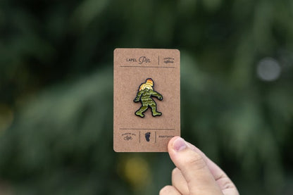 A hand holds the carded Bigfoot Adventure Pin up in front of blurred evergreen branches.