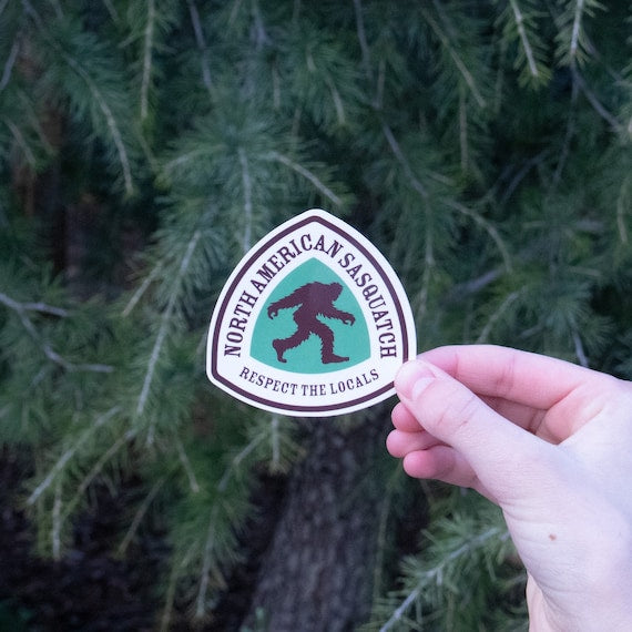 A hand holds the North American Sasquatch sticker against pine boughs, its green badge reading Respect the Locals.