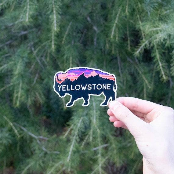 A hand holds the Yellowstone bison sticker against green pine boughs, its pink sunset sky filling the animal shape.