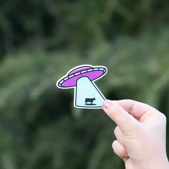 A hand holds the Holy Cow UFO Tractor Beam sticker against blurred green foliage, cow caught in the pale beam.