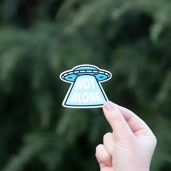 A hand holds up the Not Alone UFO sticker, its blue saucer and beam of lettering bright against dark pine.