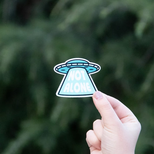 A hand holds up the Not Alone UFO sticker, its blue saucer and beam of lettering bright against dark pine.