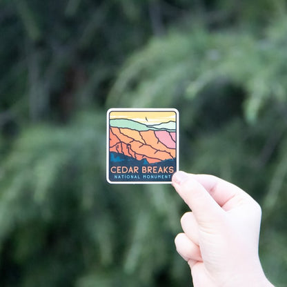 A hand holds the Cedar Breaks National Monument sticker up against blurred green conifer branches.