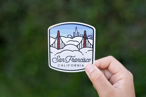 A hand holds the San Francisco California sticker against blurred green foliage, showing the bridge towers in fog.