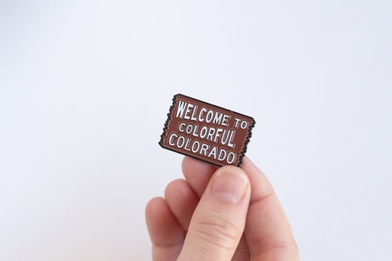 Fingers hold the brown Welcome to Colorful Colorado pin against a plain white background, white lettering facing out.