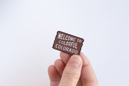 Fingers hold the brown Welcome to Colorful Colorado pin against a plain white background, white lettering facing out.