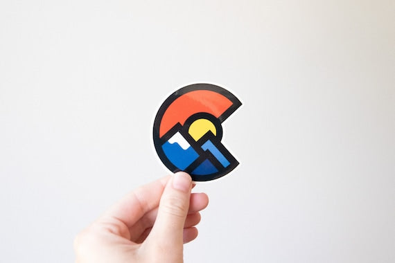 A hand holds the Colorado Mountains sticker against a plain white wall, its red C circling blue peaks and a yellow sun.