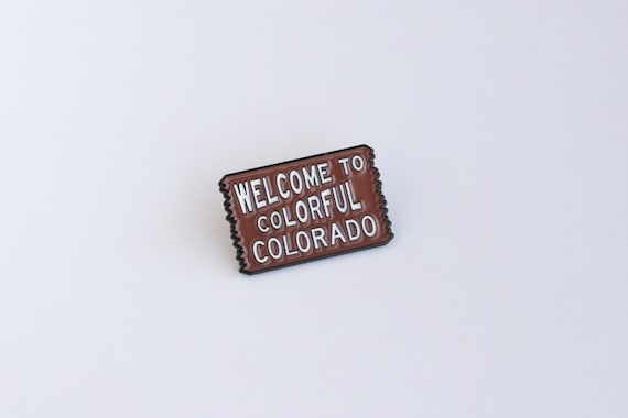 Angled on a plain white surface, the brown Welcome to Colorful Colorado pin shows its white sign lettering.
