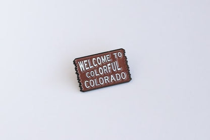 Angled on a plain white surface, the brown Welcome to Colorful Colorado pin shows its white sign lettering.