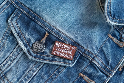 Pinned to the chest pocket of a faded denim jacket, the Welcome to Colorful Colorado pin sits beside a small charm.