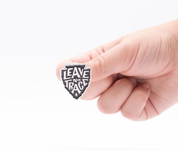 A fingertip balances the black and white Leave No Trace pin, its arrowhead shape bright against a plain backdrop.