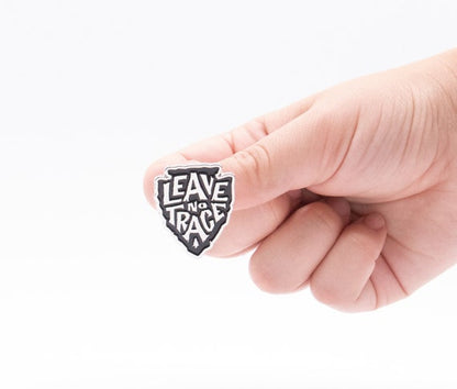 A fingertip balances the black and white Leave No Trace pin, its arrowhead shape bright against a plain backdrop.