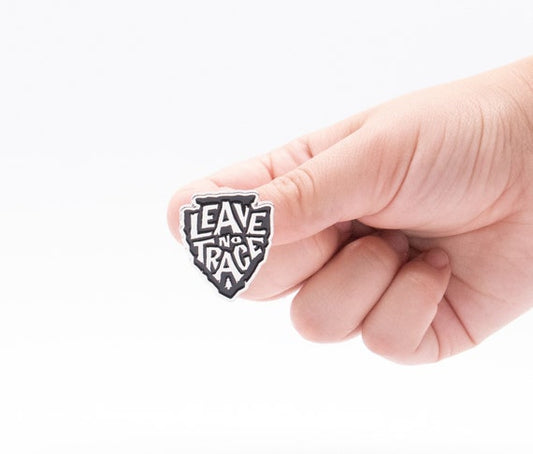 A fingertip balances the black and white Leave No Trace pin, its arrowhead shape bright against a plain backdrop.