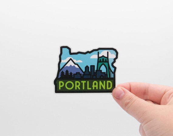 Fingers hold the state shaped Portland Oregon sticker against a plain white wall, showing the bridge and mountain art.