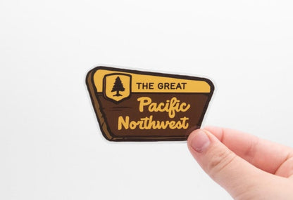Fingers hold The Great Pacific Northwest sticker against a white backdrop, its brown park sign shape and pine badge clear.
