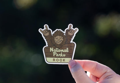 Held up against blurred dark greenery, the National Parks Rock sticker shows Sasquatch throwing two horns.