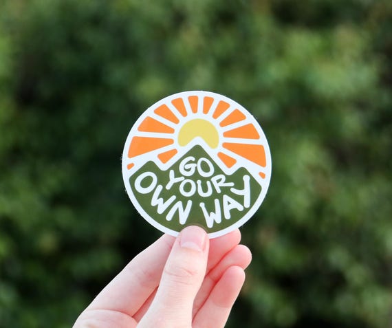 A hand holds the Go Your Own Way sticker up against a blurred green hedge.