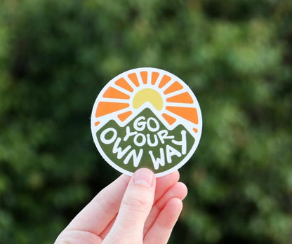 A hand holds the Go Your Own Way sticker up against a blurred green hedge.