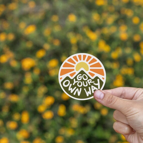 Yellow wildflowers blur behind the Go Your Own Way sticker pinched between finger and thumb.