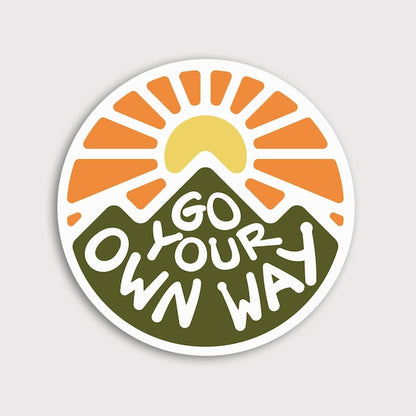 Round Go Your Own Way sticker on a pale grey background, orange sun rising behind a green mountain.
