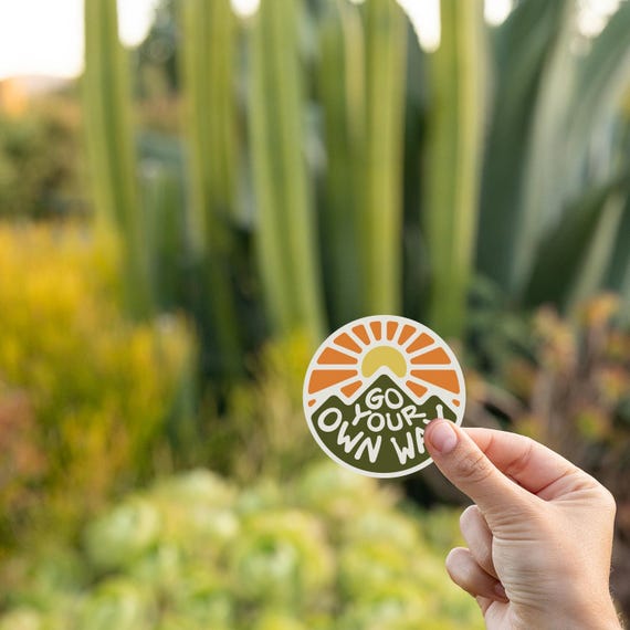 Fingers hold the Go Your Own Way sticker in front of tall cactus and golden garden plants at dusk.