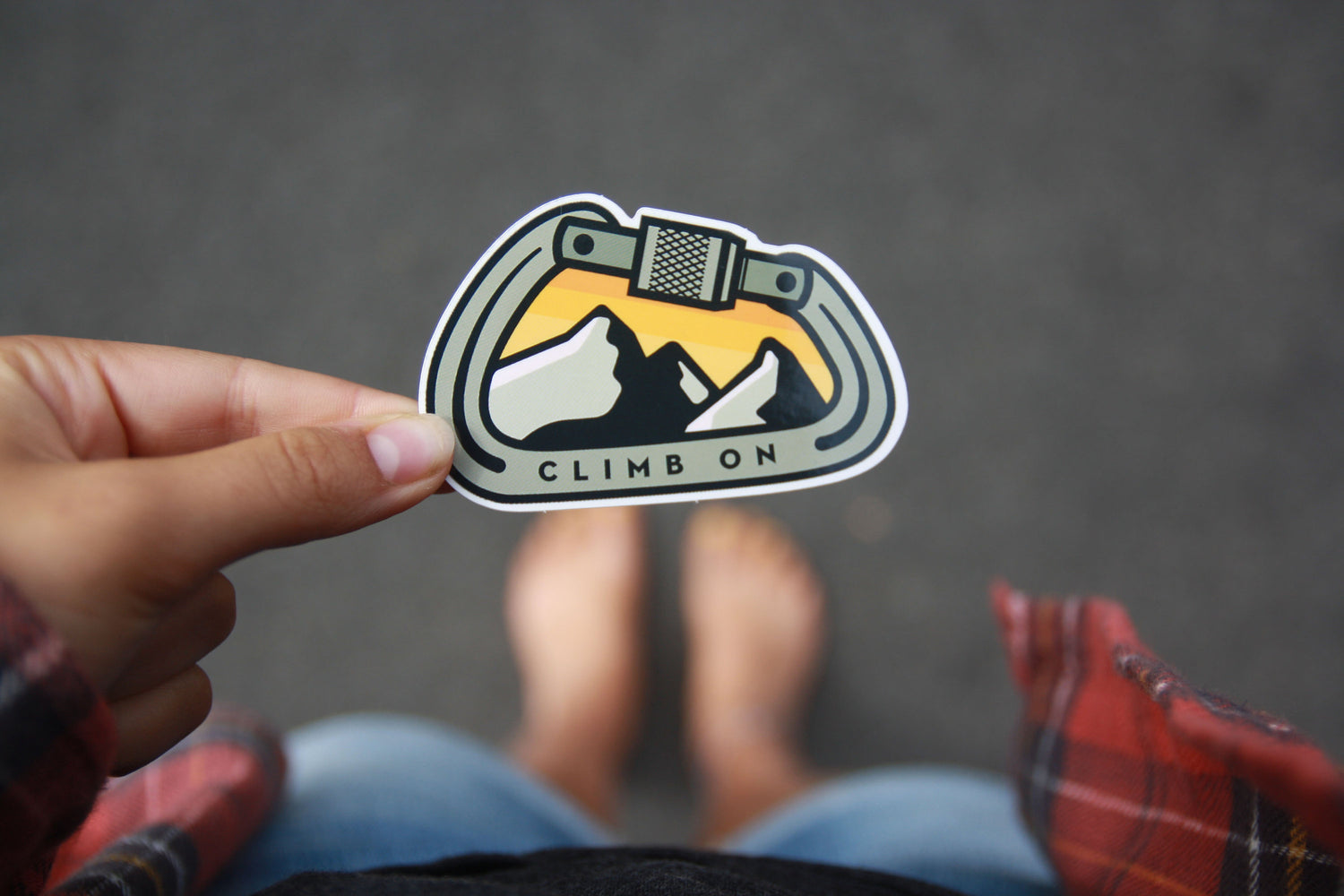 Looking down past bare feet and plaid, a hand holds the Climb On Carabiner sticker over grey asphalt.