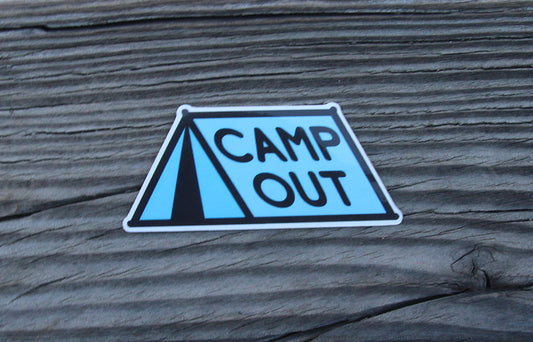 Resting on weathered grey wood, the Camp Out sticker shows its black tent outline and blue panels.