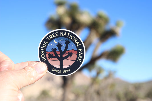 Blue desert sky and a real Joshua tree stand behind the Joshua Tree National Park sticker held up in fingers.