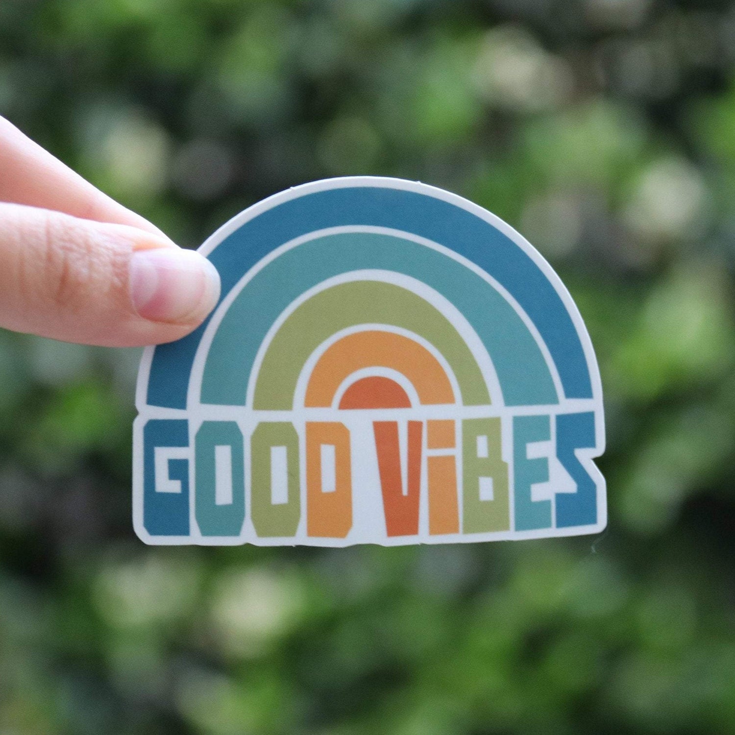 A fingertip steadies the Good Vibes sticker close to the lens, its rainbow arcs bright against blurred green leaves.