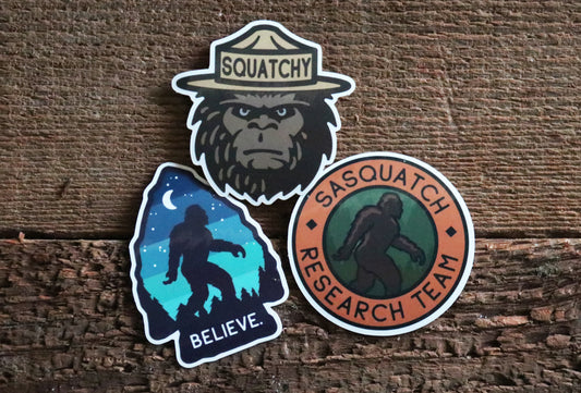 Three stickers rest on rough bark, with the Sasquatch Research Team circle beside the Squatchy face and Believe designs.