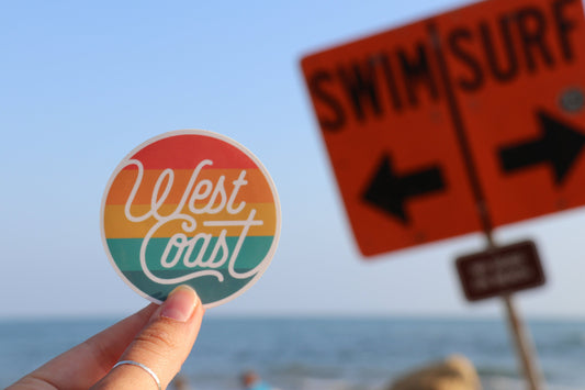 Beach light behind, the West Coast Life sticker is raised near an orange SWIM SURF sign above the ocean.