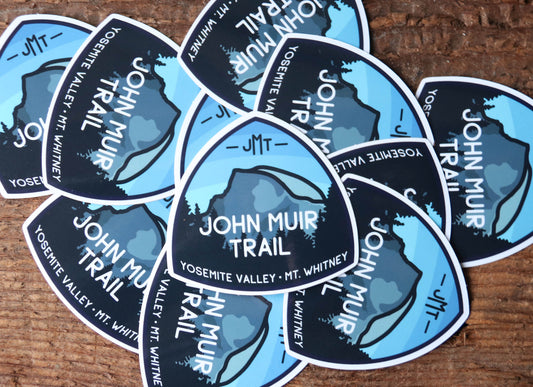 Scattered across weathered wood, a pile of John Muir Trail stickers overlaps face up and sideways.