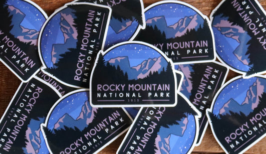 Scattered across dark wood planks, a pile of Rocky Mountain National Park stickers shows starry peaks and pines.