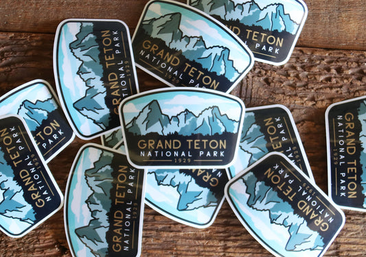 A scattered pile of Grand Teton National Park stickers lies across weathered grey wood planks.
