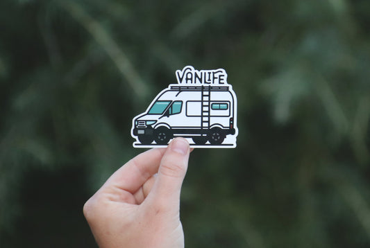 Held further back against dark conifer branches, the white Vanlife Sprinter sticker shows its roof ladder.